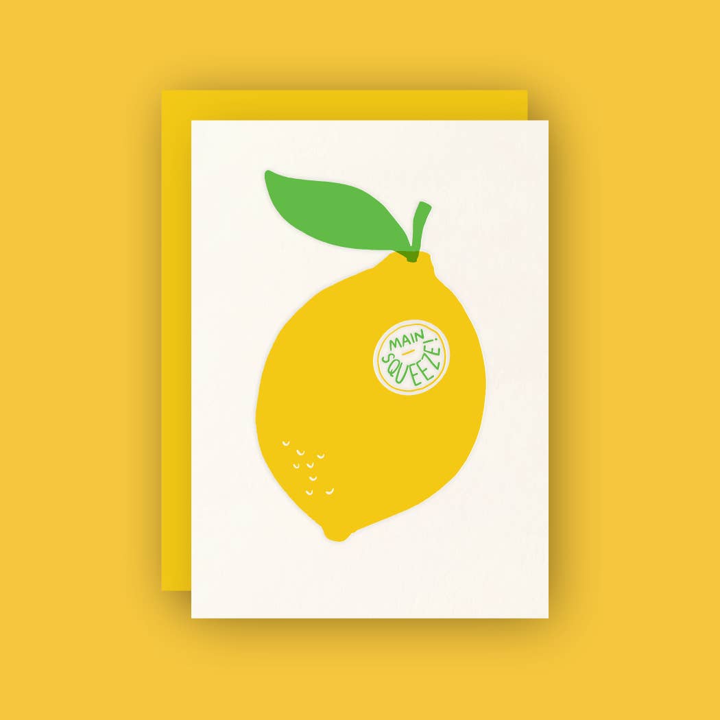 A white greeting card features a large, bright yellow lemon with a green leaf on top and a circular sticker reading "MAIN SQUEEZE" in green text. The card is placed on a matching yellow envelope against a solid yellow background.