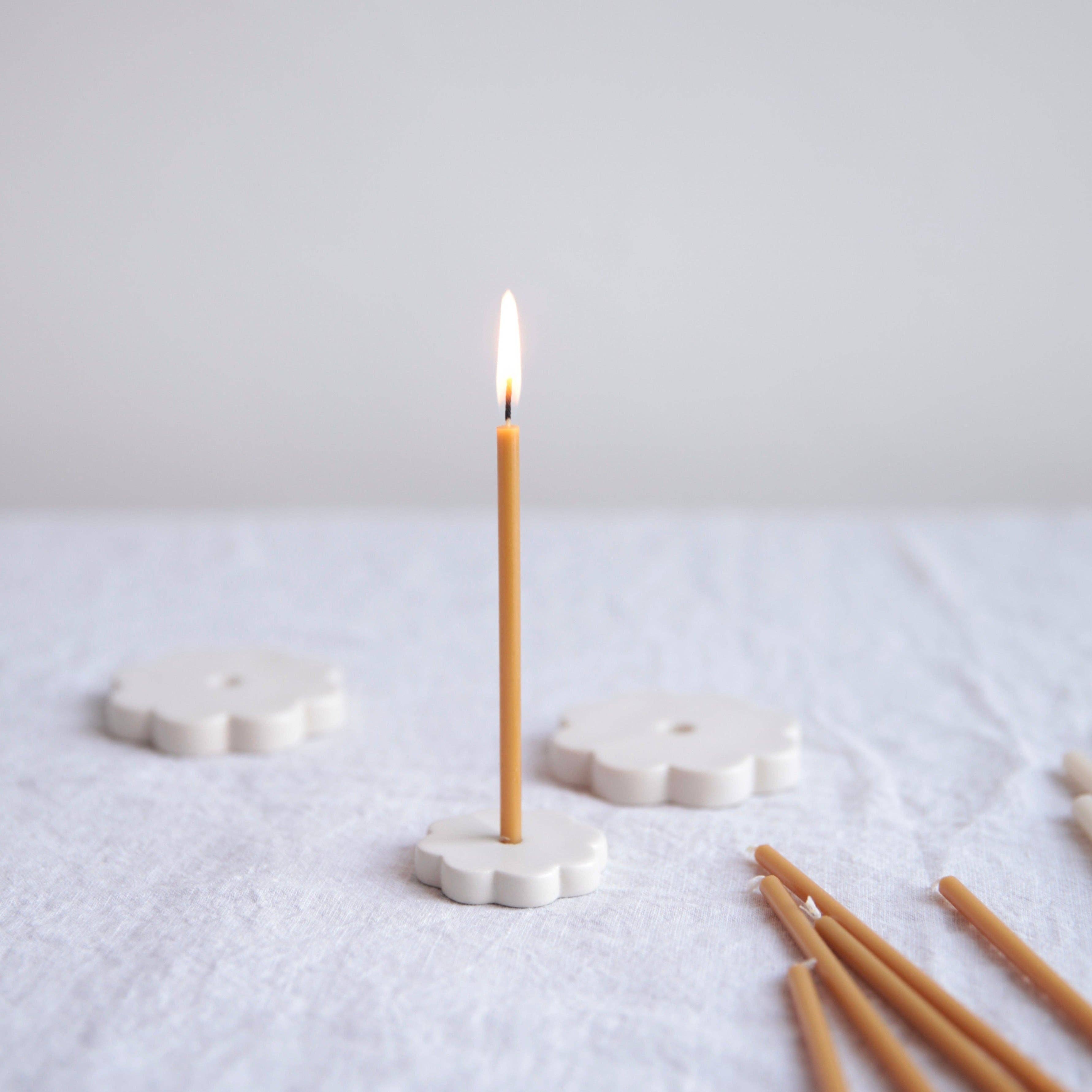 A single lit taper candle stands upright in a white flower-shaped holder on a textured white fabric surface, with additional unlit taper candles and more holders softly blurred in the background.