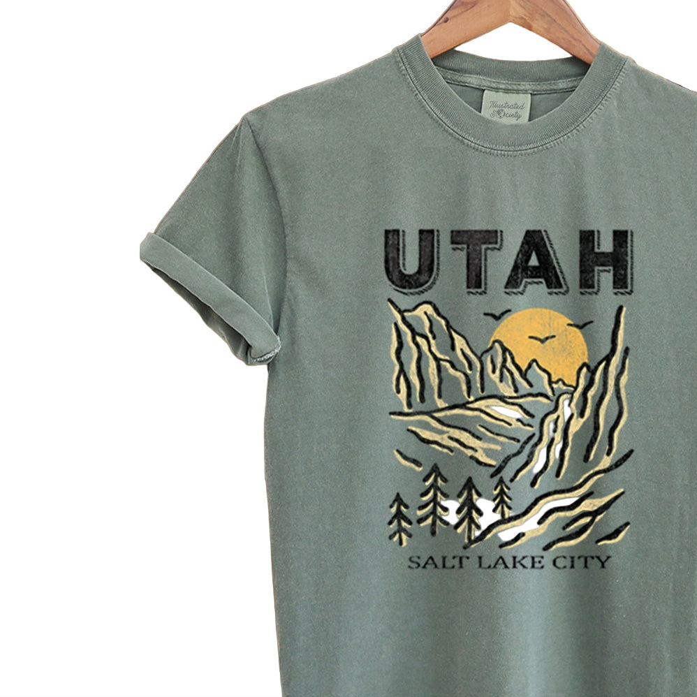 Green t-shirt with 'UTAH' and mountain graphic on a hanger against a textured background