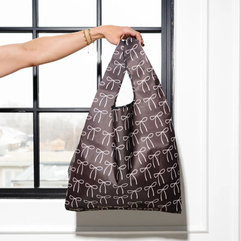 A person is holding up a reusable shopping bag by its handles in front of a window. The bag is dark brown with a repeating pattern of cream colored bow illustrations. 