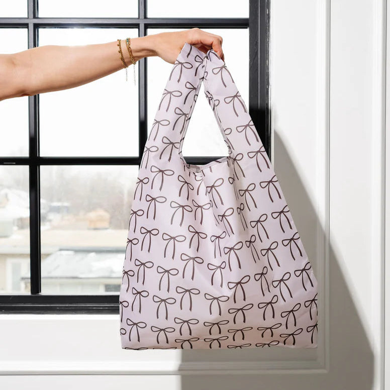 A person is holding up a reusable shopping bag by its handles in front of a window. The bag is cream colored with a repeating pattern of brown bow illustrations.