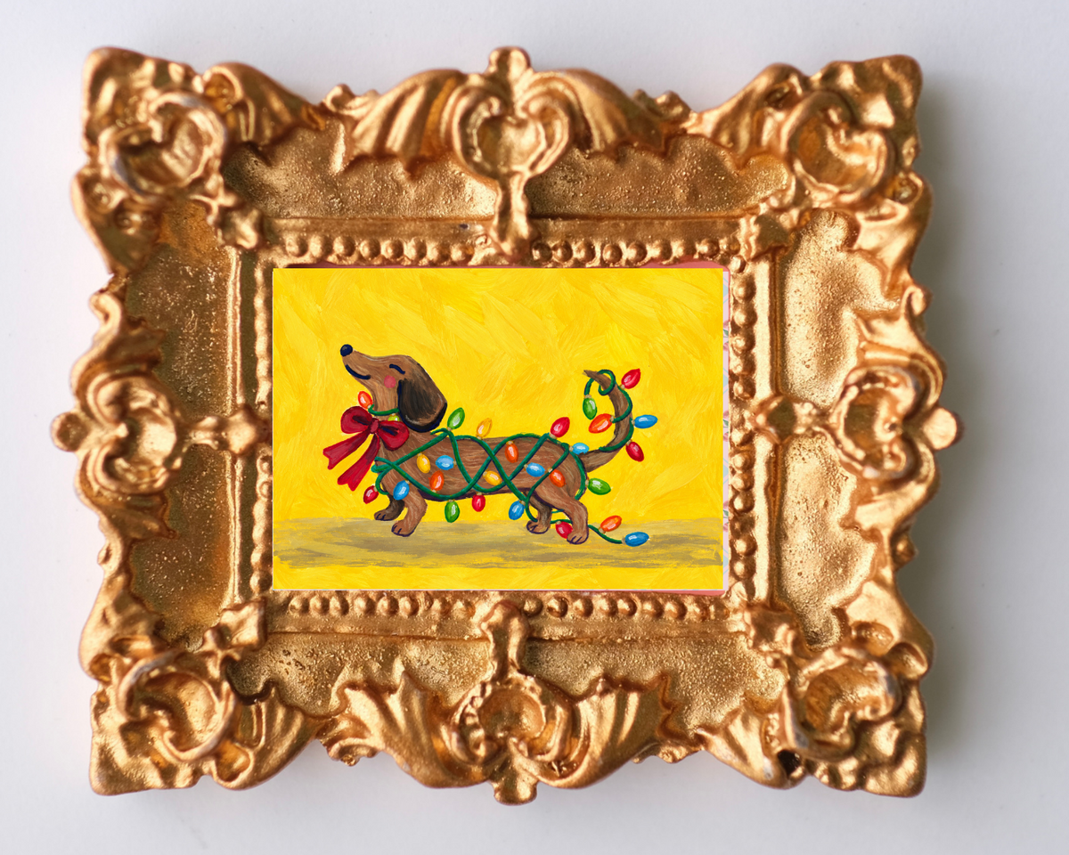 Ornate gold-framed Christmas magnet featuring a playful dachshund tangled in colorful Christmas lights with a red bow against a bright yellow background.