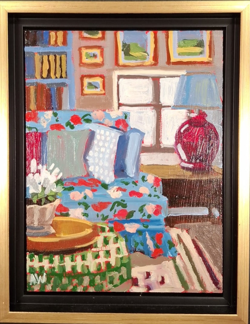 A scene from a living room with a blue couch that has a floral pattern. There are big pillows with patterns on the couch. There is a green plaid ottoman in the foreground with a gold tray holding a vase of flowers. Next to the couch is a brown side table with a red lamp. On the wall behind it all is a blue bookshelf full of books and many pieces of art on the wall, and the shadow of the window lighting up the remaining wall space. 