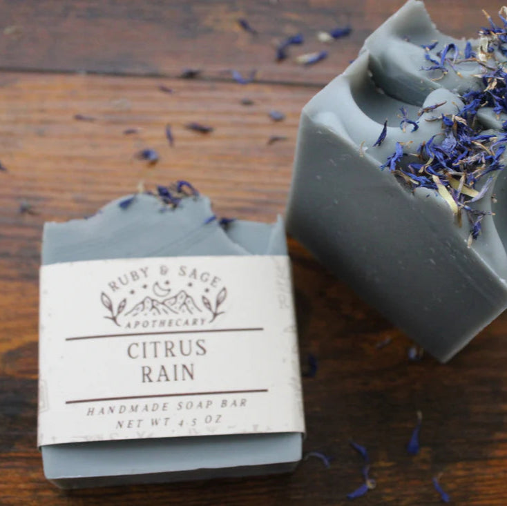 A handmade gray soap bar labeled "Citrus Rain" from Ruby & Sage Apothecary sits on a wooden surface, with another bar beside it topped with dried purple and white flower petals. The soap has a smooth, sculpted top and a natural, artisanal appearance.
