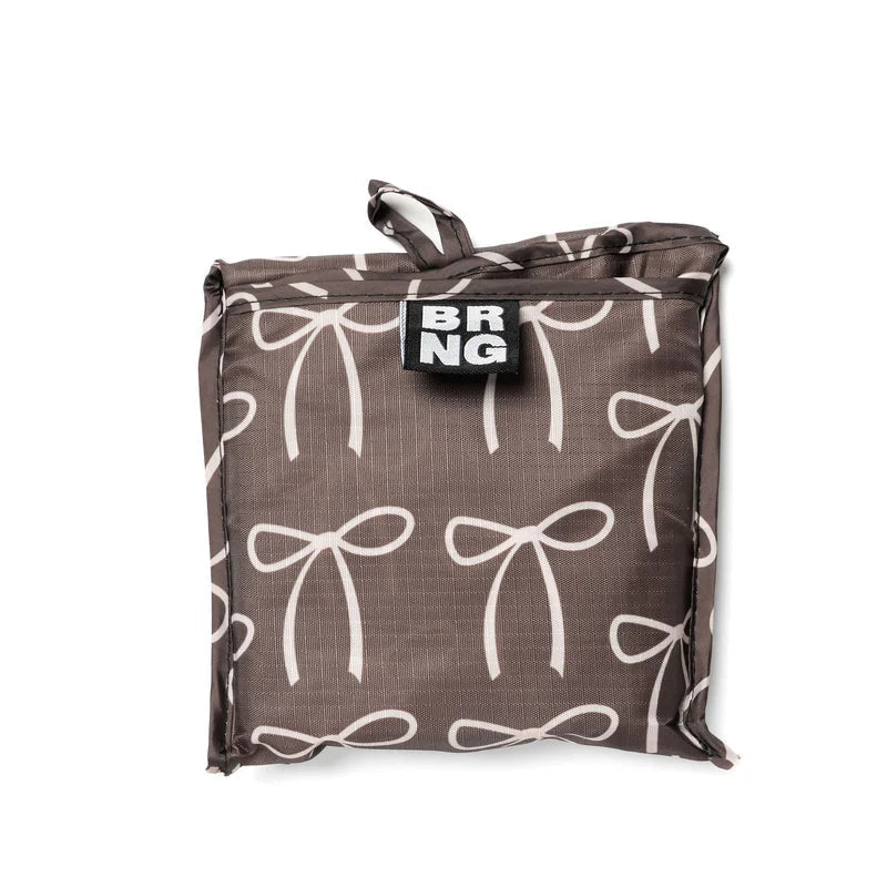 This image shows a compact, folded reusable shopping bag with a dark brown background and a pattern of cream colored bows. The bag is neatly packed into a square shape with a small fabric loop at the top and a "BRNG" logo label sewn on the front.