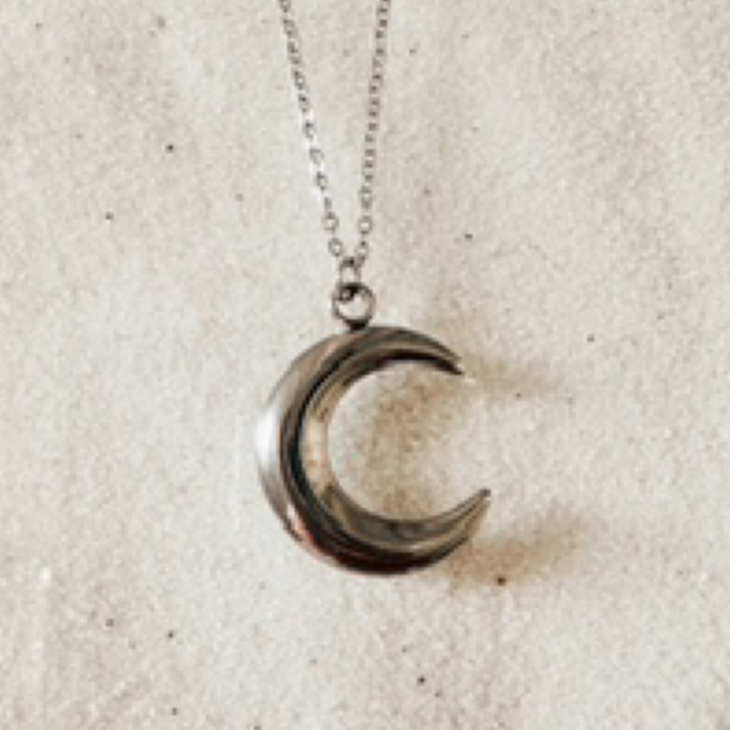 Delicate silver chain with large silver crescent moon charm. Pictured against white sand.