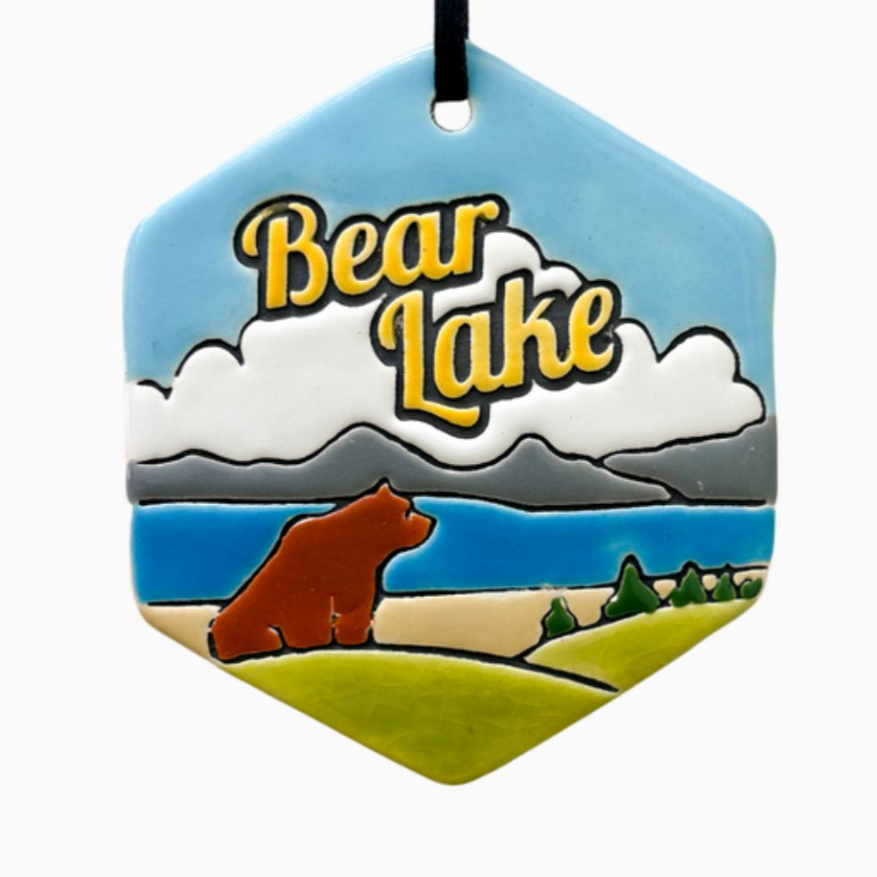 Hexagon-shaped ceramic ornament featuring a scenic landscape with a brown bear near a lake, green hills, pine trees, and distant mountains under a blue sky. The words “Bear Lake” are prominently displayed in bold yellow letters across white clouds.