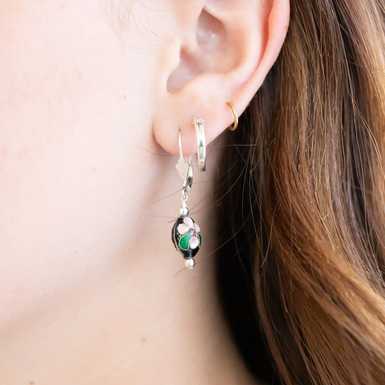 Silver clasp earring with a black oval bead that has a pink painted flower. Pictured on a brunette girls ear with other minimalistic hoops.
