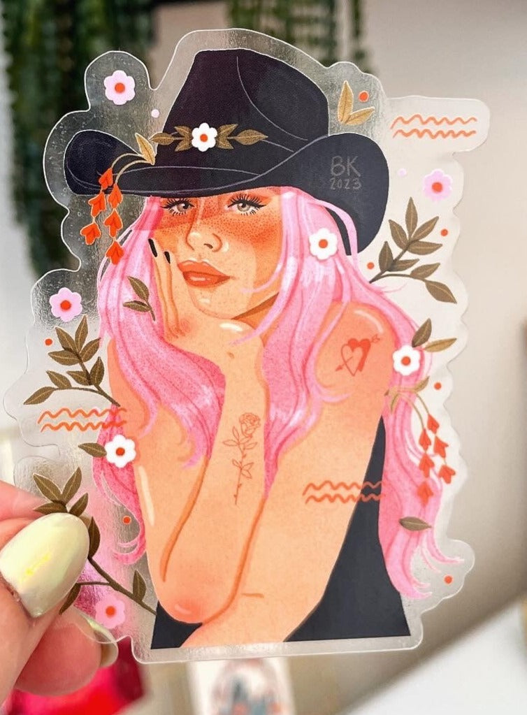 Cowgirl Sticker. A cowgirl with long pink hair wearing a cowgirl hat rests her head in her palm. She has a tattoo of a heart with an arrow in it and is surrounded by florals. 