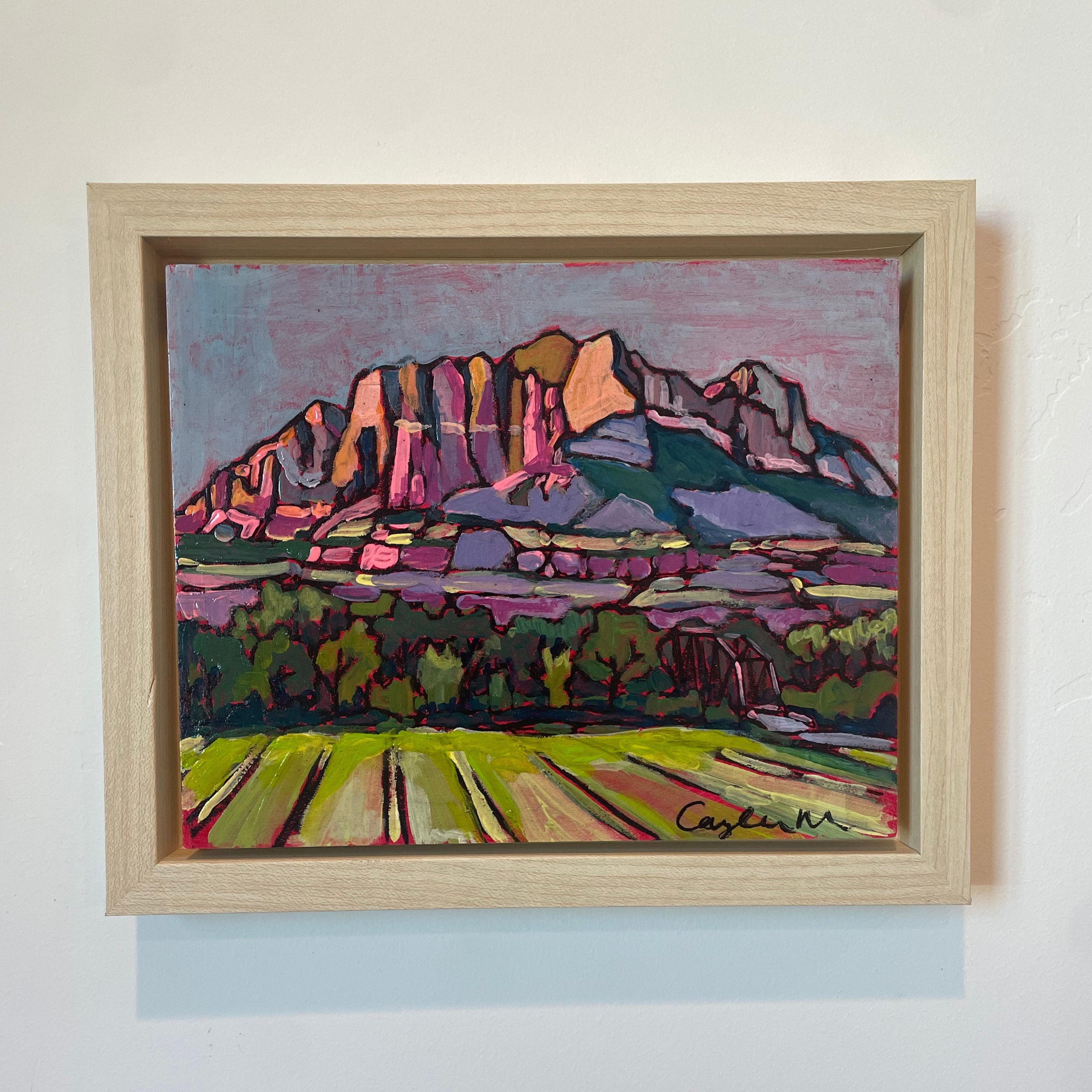 A view of the rocky Mt. Kinesava at dusk. Bright hues of pink, purple, orange, green, and blue are used to give a moody dusk feeling. The field in the foreground is bright green with leafy trees behind it. The sky is blue and purple, 