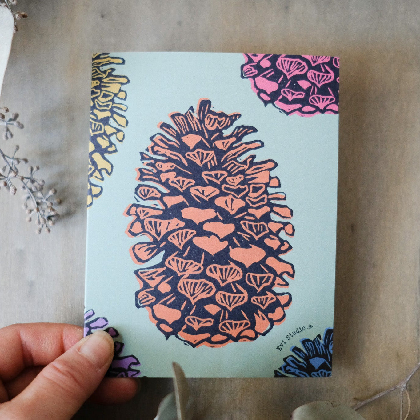 Light sage green card with a large brown pinecone centered in the middle, other colorful pinecones are cut off along the edges, showing just partial images. Other pinecones are pink, yellow, blue, and purple. Pictured against a wooden background in a woman's hand.