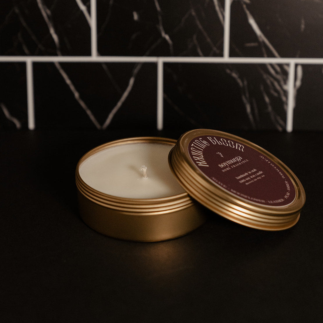 Small gold tin candle with a burgundy label. Pictured against a black marble back splash.