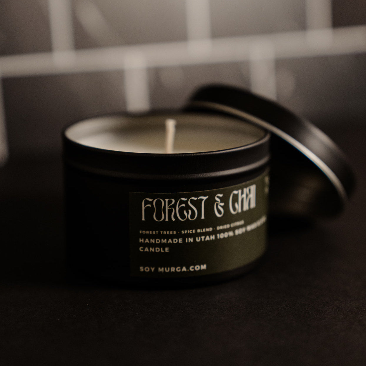 Small black matte candle with a moss green label and lid off. Pictured against a black marble back splash. 