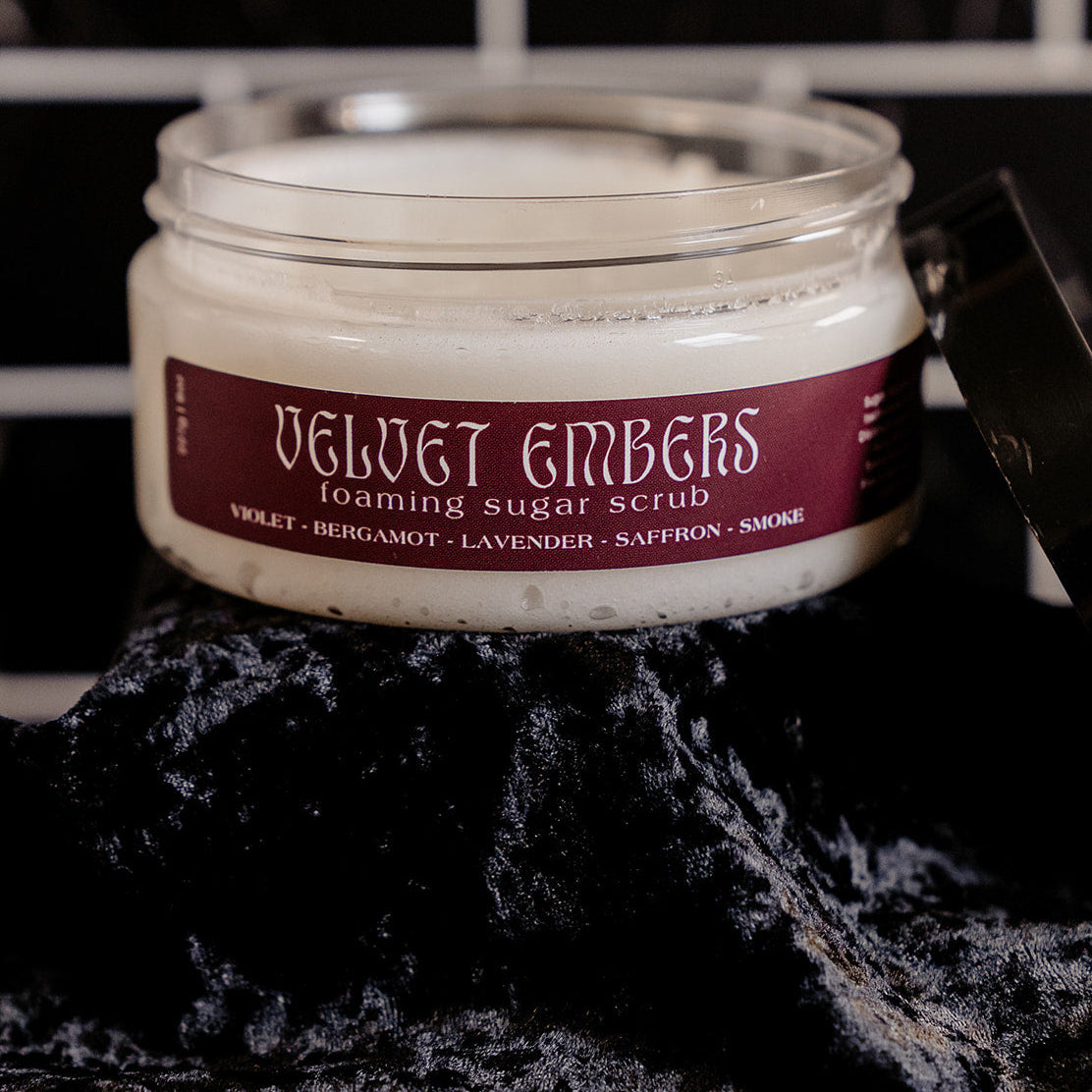 Small shallow container with a burgundy label with lid off and leaning against containers side. Pictured with black velvet and a black marble backsplash. 