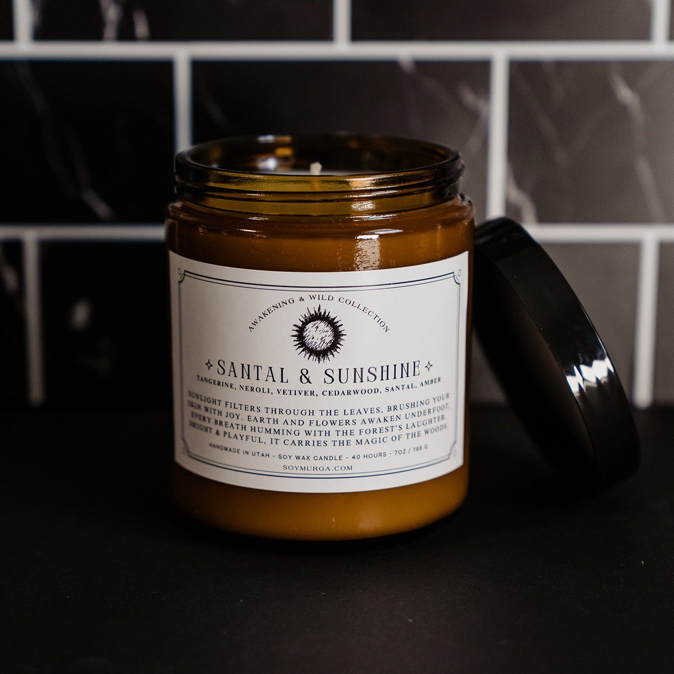 Santal & Sunshine candle in an amber glass jar on a black countertop with black marble subway tile in the background. A custom label adhered to the front of the label. 