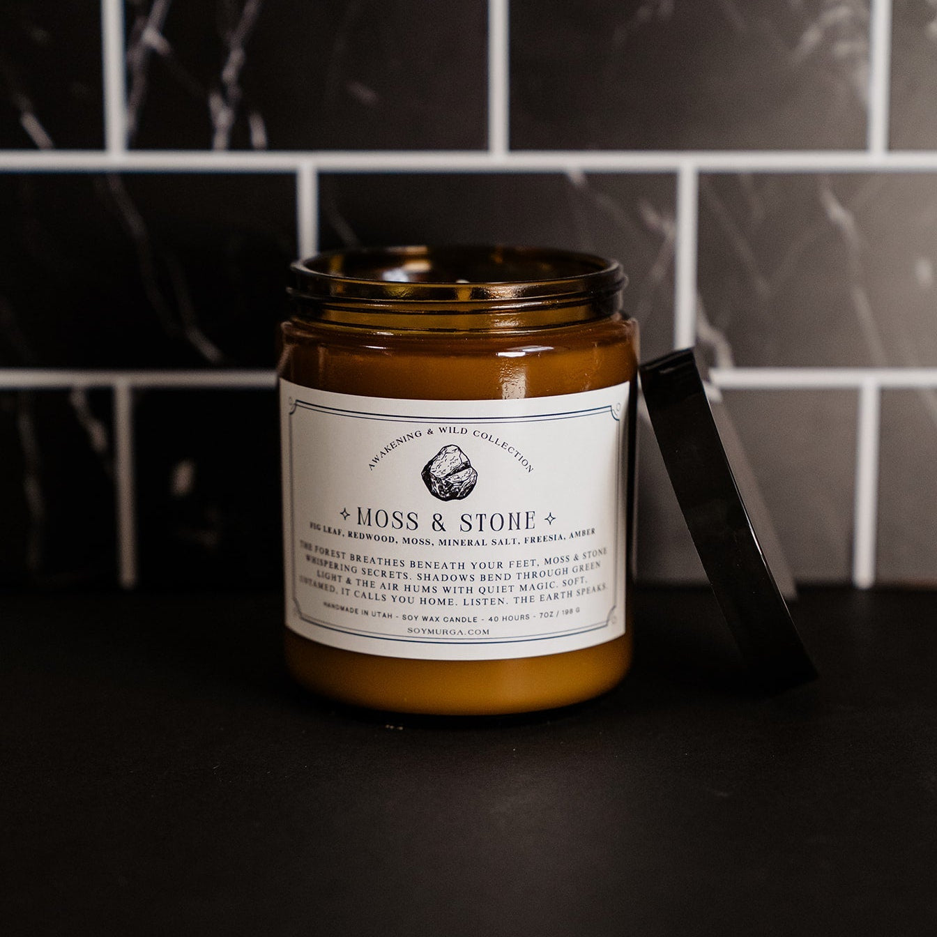 The moss & stone candle in an amber glass jar with a custom label on the front. Candle is on a black countertop with marble black subway tile in the background.