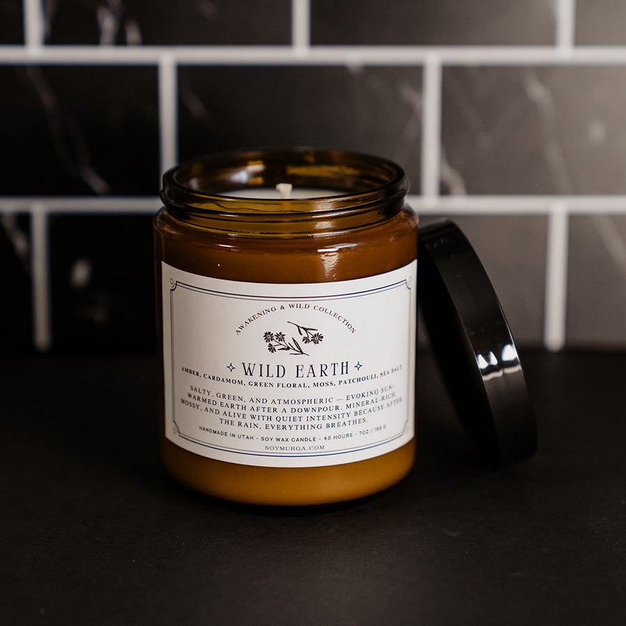 The Wild Earth candle on a black countertop with black marble subway tile in the background. The candle is in an amber jar with a custom label printed on the front. 