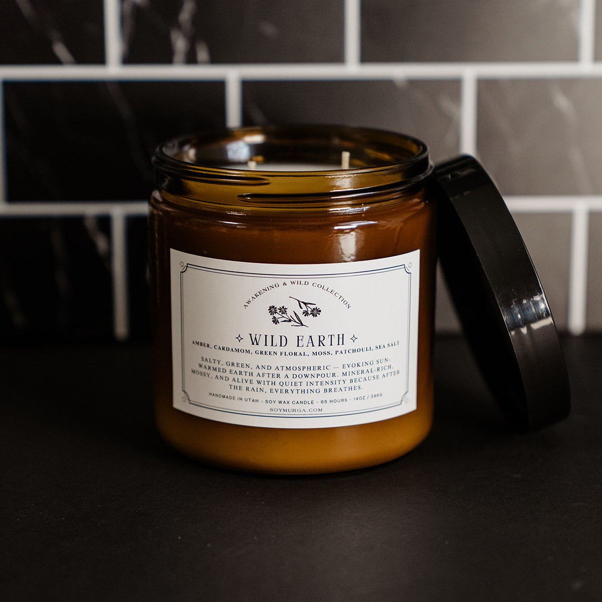 14 oz Wild Earth candle in an amber glass jar with two wicks on a black countertop with black marble subway tile in the background. 