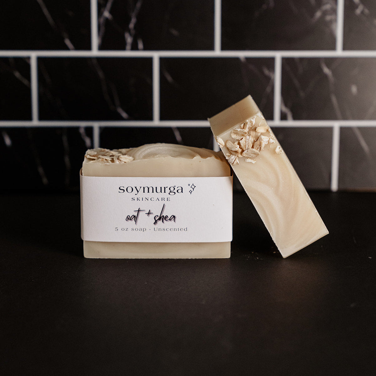 Oat + Shea bar of soap on a black countertop with black marble subway tile in the background. 