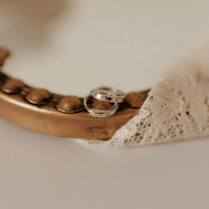Two silver rings with small inset diamonds rest on a vintage-style, bronze-beaded surface partially draped in delicate ivory lace. The rings feature a modern, minimalist design with evenly spaced stones, creating an elegant contrast against the antique background.