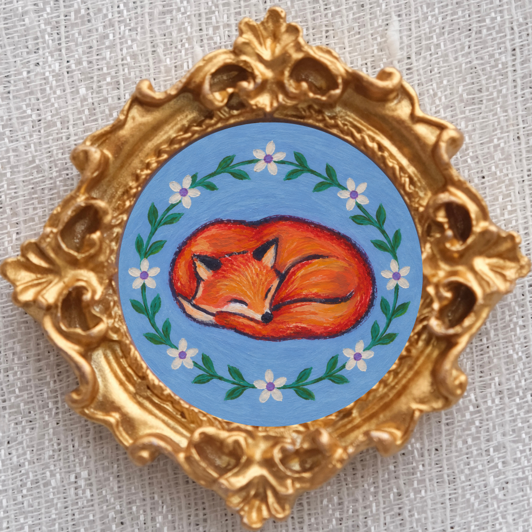 Tiny red fox curls up in a ring of flowers inside a tiny ornate gold frame. 