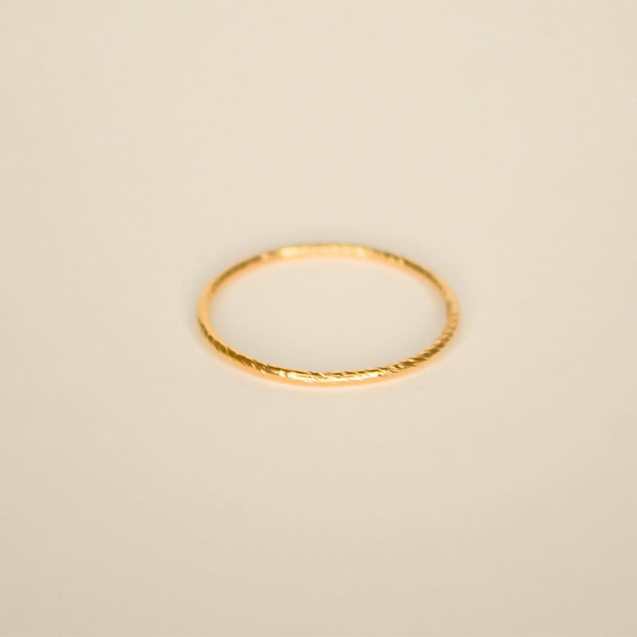 Thin gold minimalist band with intricate texture that catches the light pictured on an off white back ground.