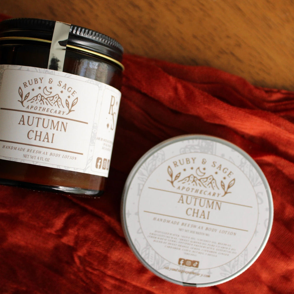 A small tin and glass jar of Autumn Chai body lotion. Pictured against a rust colored table runner. 