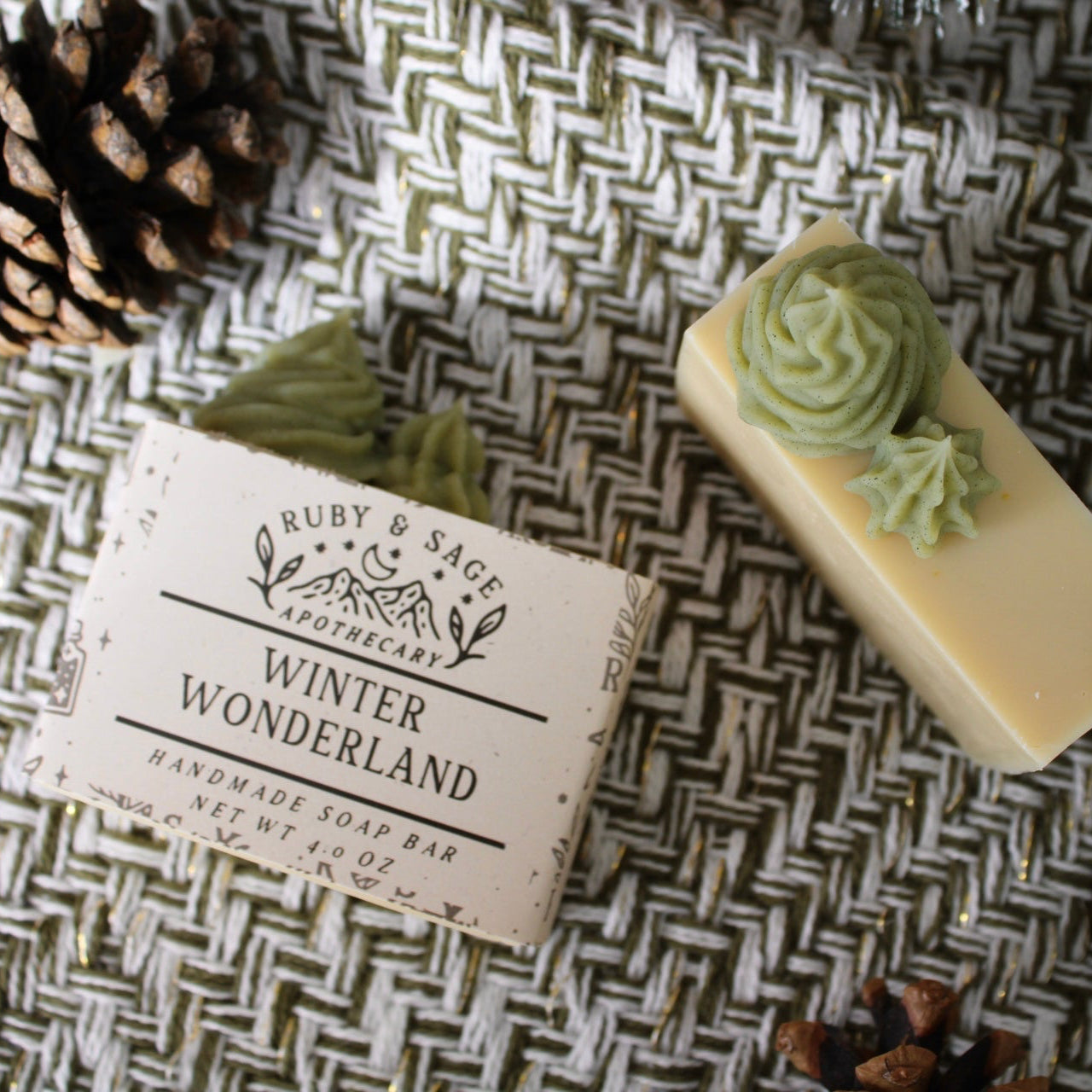 Cream soap bar with light green piped trees on top. Labeled with cream paper, designed with sprigs and various fonts. Pictured against a white and green weave with pinecones and frosted pines.
