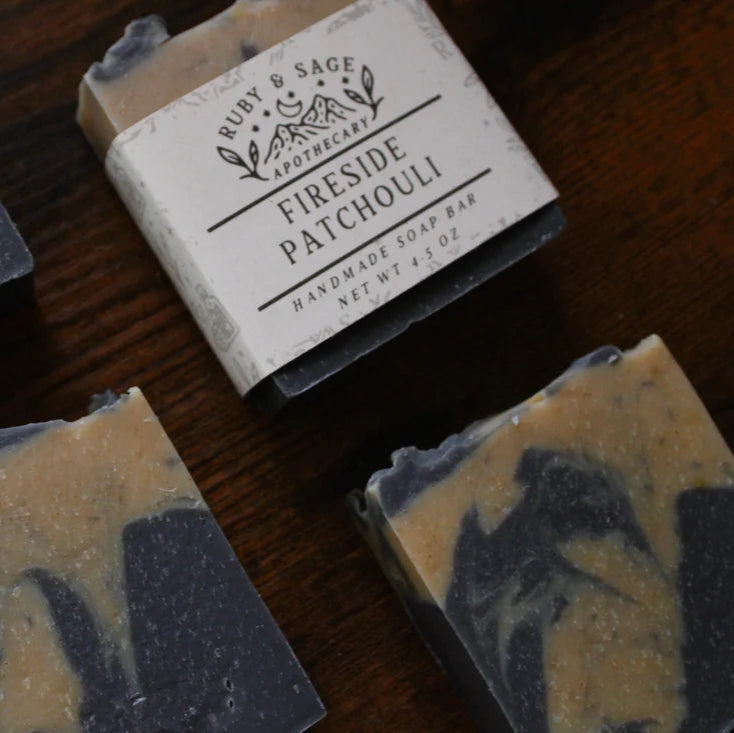 A collection of handmade soap bars from Ruby & Sage Apothecary is arranged on a wooden surface, with one bar prominently labeled “Fireside Patchouli.” Each bar features a two-tone marble design with brown and black swirls, reflecting the natural, nourishing ingredients.