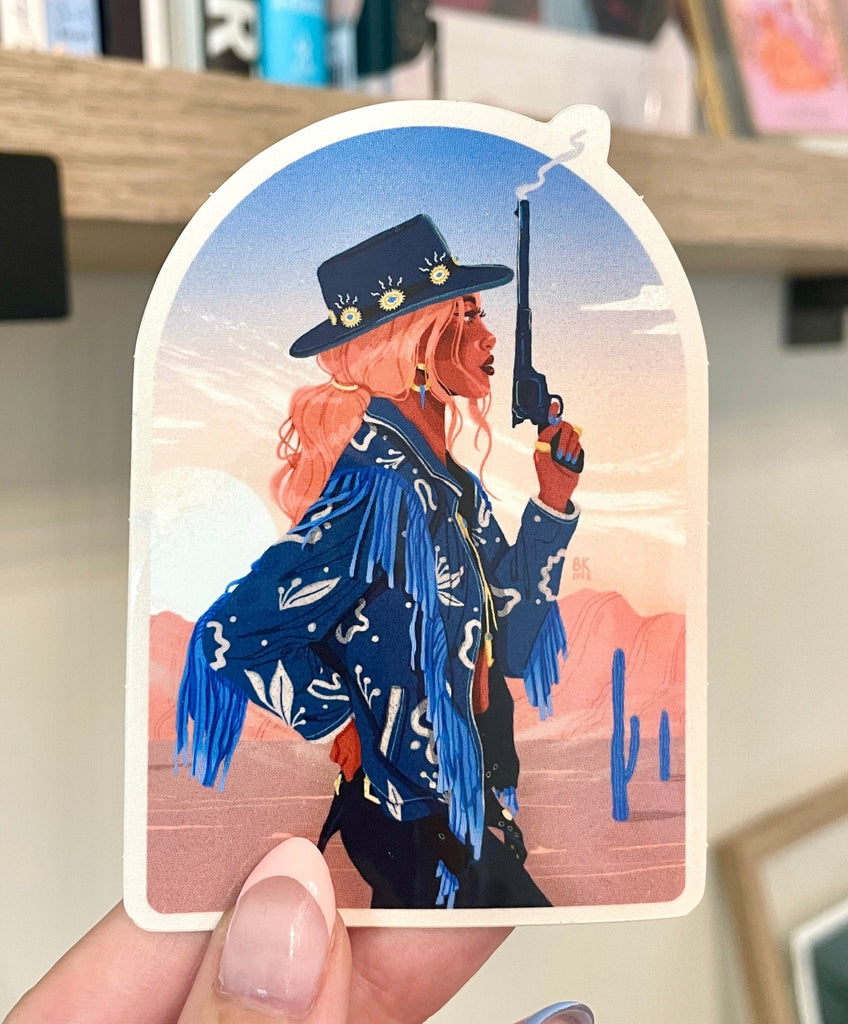 An arched vinyl sticker of Gunsmoke. A cowgirl, wearing a cowgirl hat and a denim jacket with fringe, holds a smoking gun pointed towards the sky. She faces east so we can only see her side profile. She has her pink hair back in a low ponytail and she is standing in a desert with red rocks, cacti, and blue sky. 