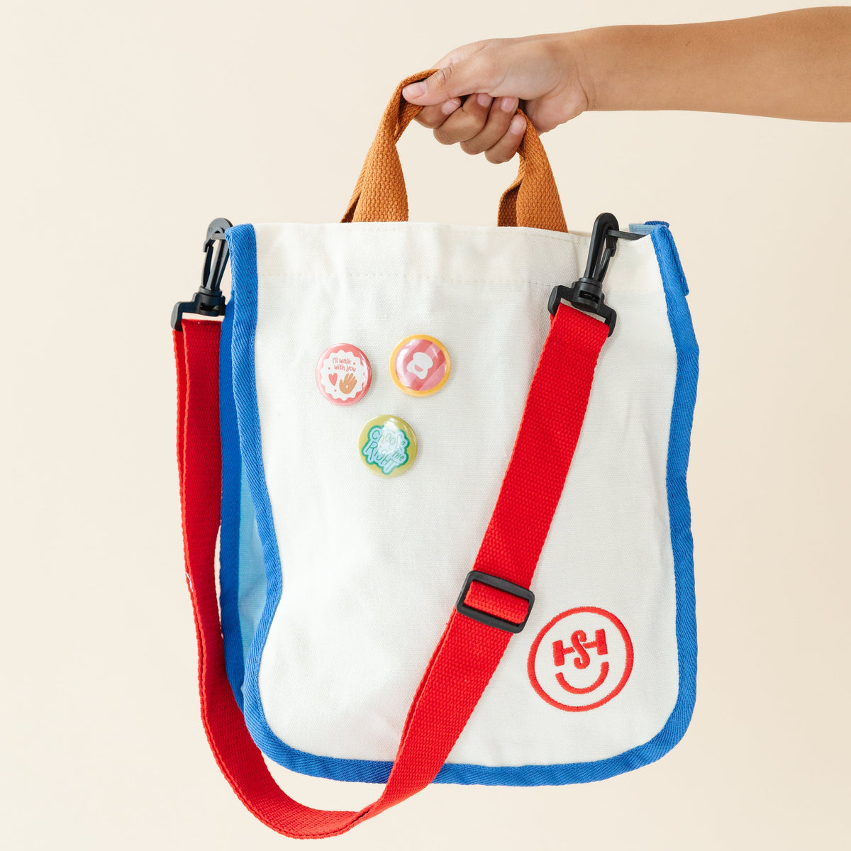 A cream canvas tote bag with blue trim, orange handles, and a red adjustable shoulder strap is held by a hand against a light background. The bag features three colorful decorative pins and a red circular “HS” smiley logo. 