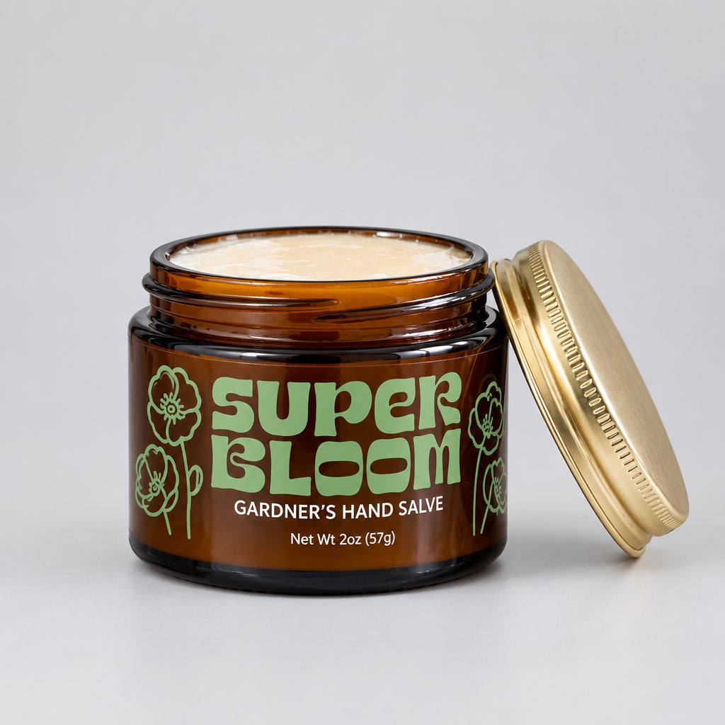 Super Bloom Gardener’s Hand Salve in an amber jar with the lid beside it, displayed on a marble surface among potted greenery.