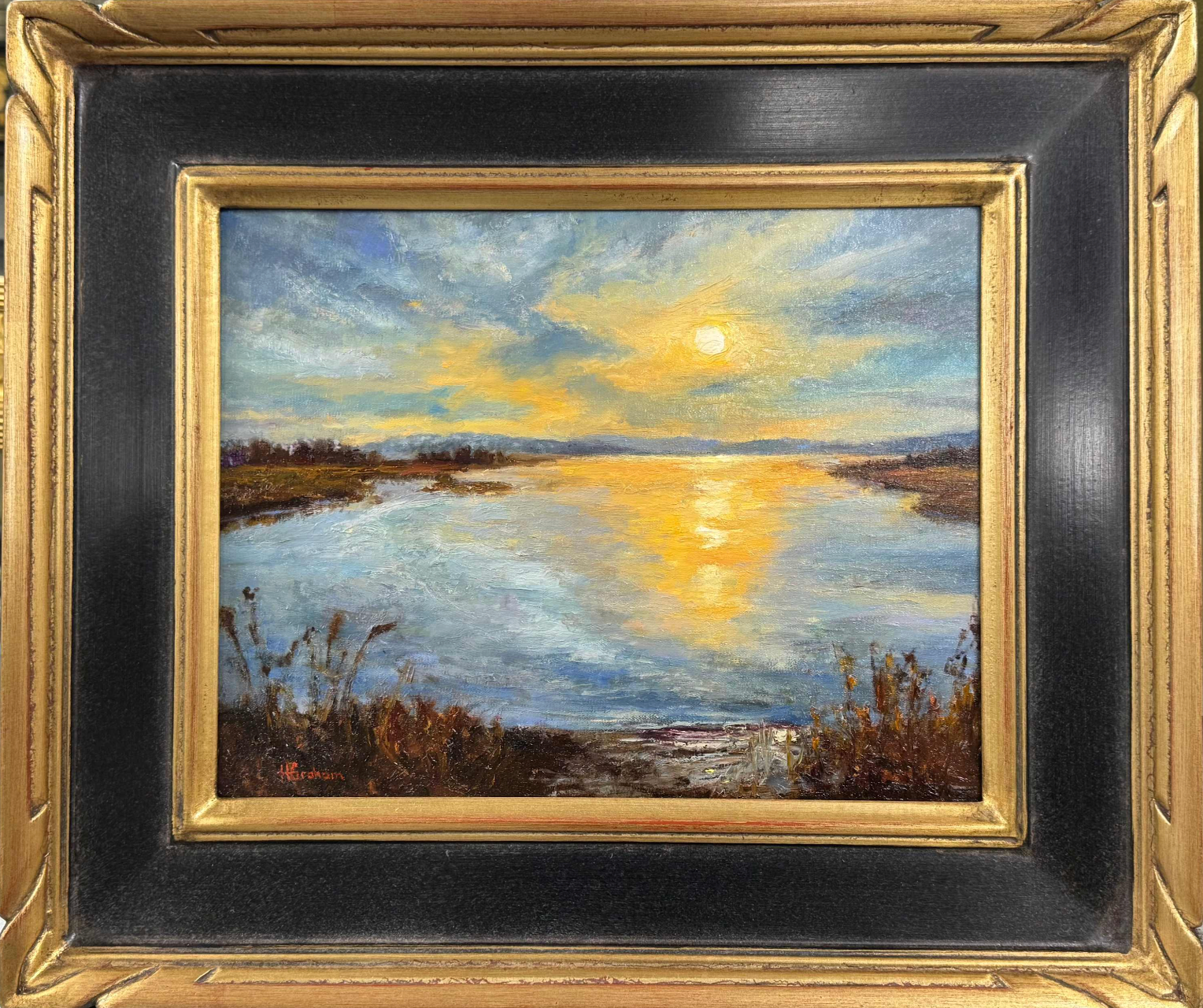 A beautiful water landscape surrounded by tall grasses and flora, with the sun about to set and the orange and yellow hues reflected in the water. The sky is partly cloudy and there are mountains in the distant background. 