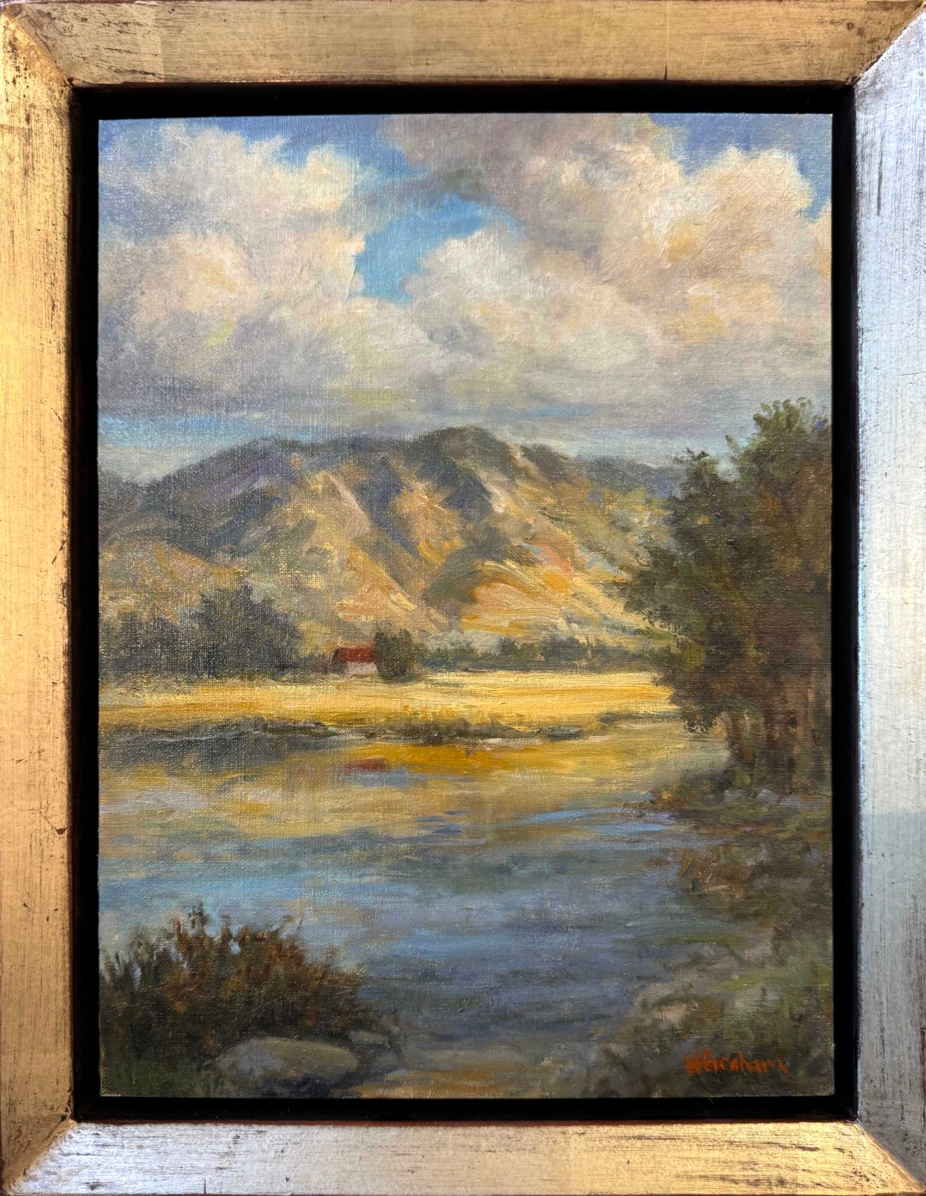 A view of a shoreline with a wide river and golden grasses on the shoreline and a red and white barn in the background. Tall mountains colored in gold and orange as the sun sets opposite from them. The sky is blue and full of fluffy white clouds. 