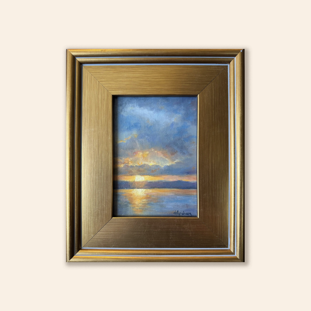 An image of the sun halfway set over water with a mountain range in the scene. Blue and yellow clouds fill the sky as dusk begins to set and the sun is reflected in orange and yellow on the water. Framed in a thick gold frame. 