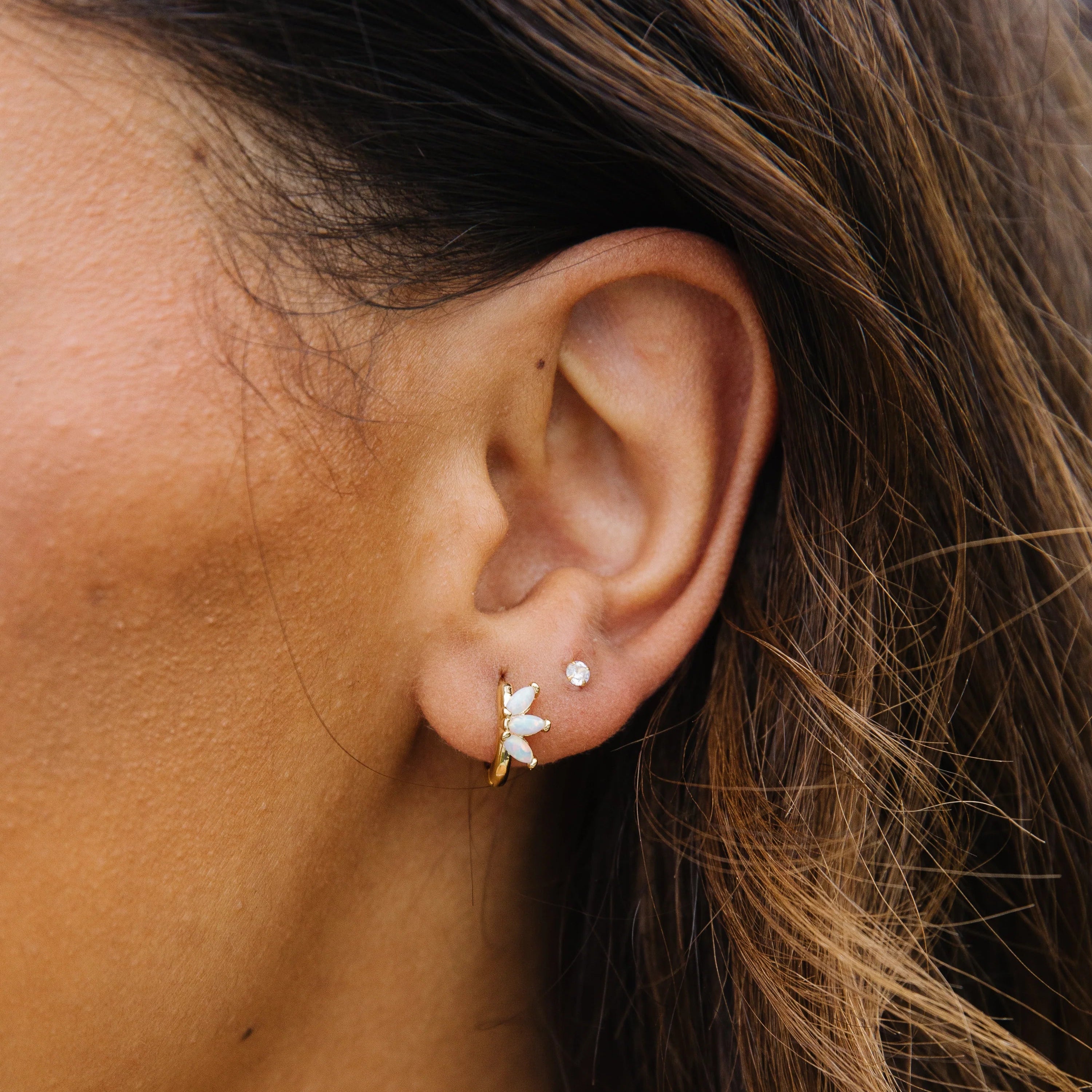 This image showcases a close-up of a woman wearing delicate opal and gold huggie earrings, paired with a small crystal stud in the second piercing. The primary earring features a trio of marquise-shaped opals arranged in a fan or floral design, giving it a soft, feminine aesthetic with a subtle shimmer.