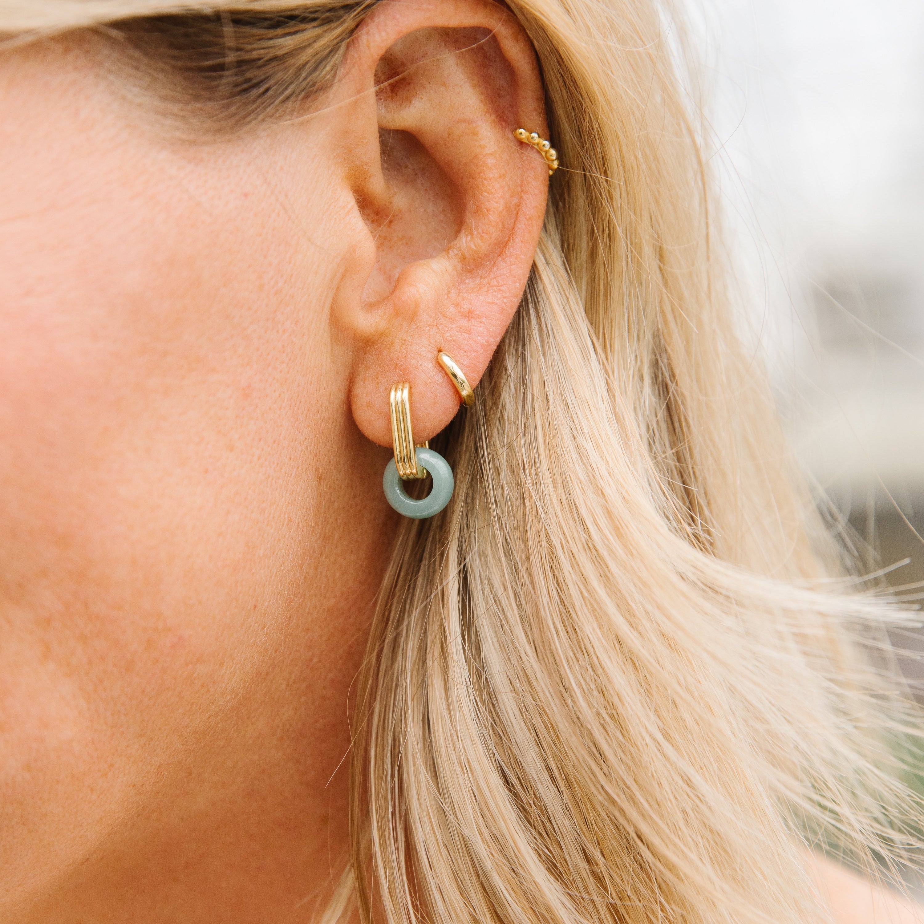 Gold ribbed rectangle huggies with a mini donut jade charm looping through the huggie. Pictured on a blonde woman's ear with other minimalistic gold huggies. 
