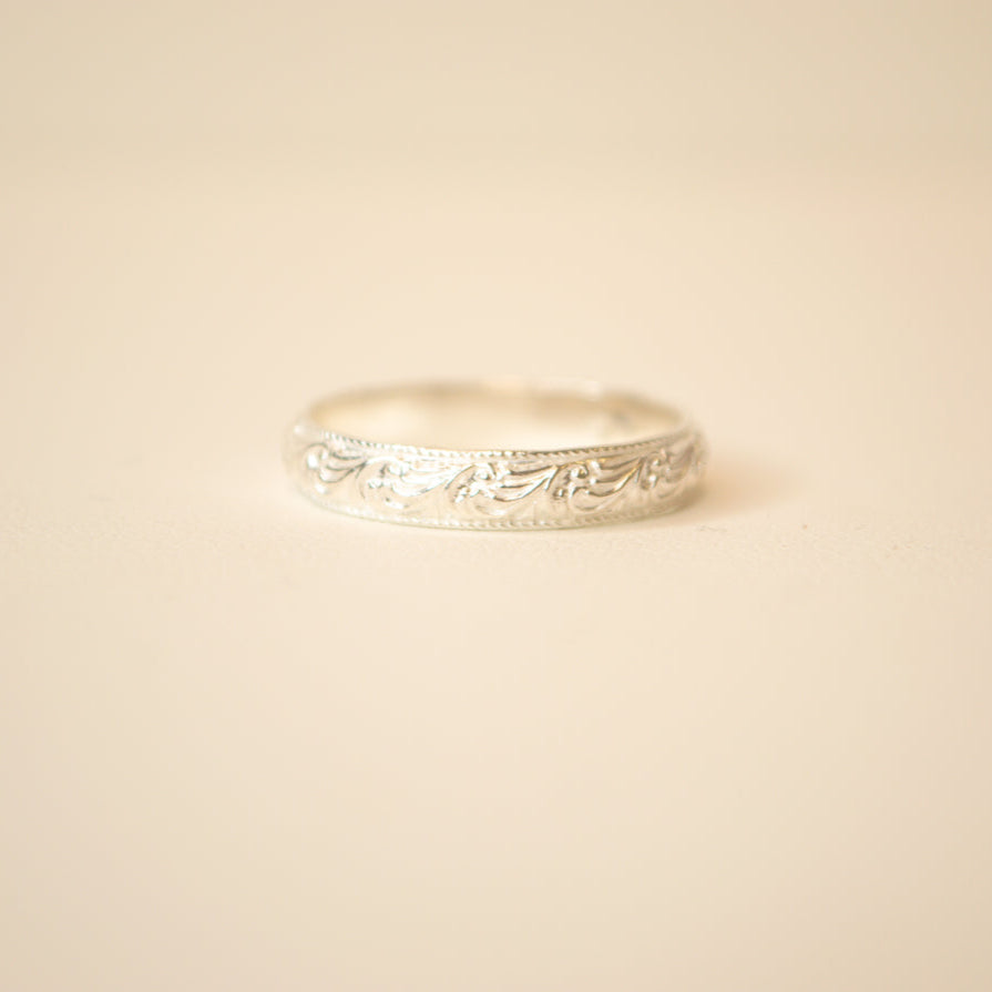 Silver band with delicate floral filagree engravings on an off white background.
