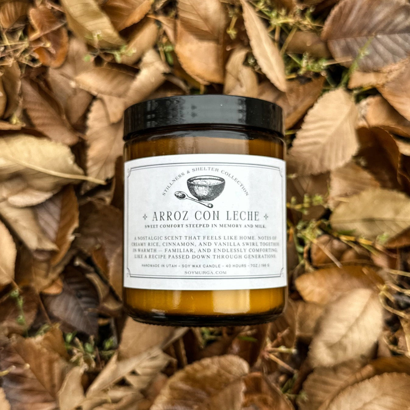 Arroz Con Leche candle in an amber glass jar lying in a pile of fall leaves. A custom label is adhered to the front. 