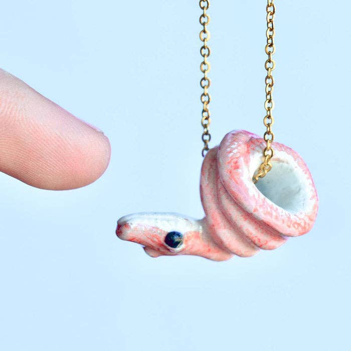 Add a pop of color and a dash of daring with our Pink Python Necklace! This porcelain pendant is hand-painted in perfect pink hues, capturing the allure of this fascinating reptile. Hung from a delicate gold chain, it's a bold yet elegant piece that's sure to catch eyes. Due to the handmade nature of this product, slight variations in color and design may occur, making each piece truly one-of-a-kind. Perfect for reptile enthusiasts, lovers of unique jewelry, or anyone looking to make a vibrant statement.