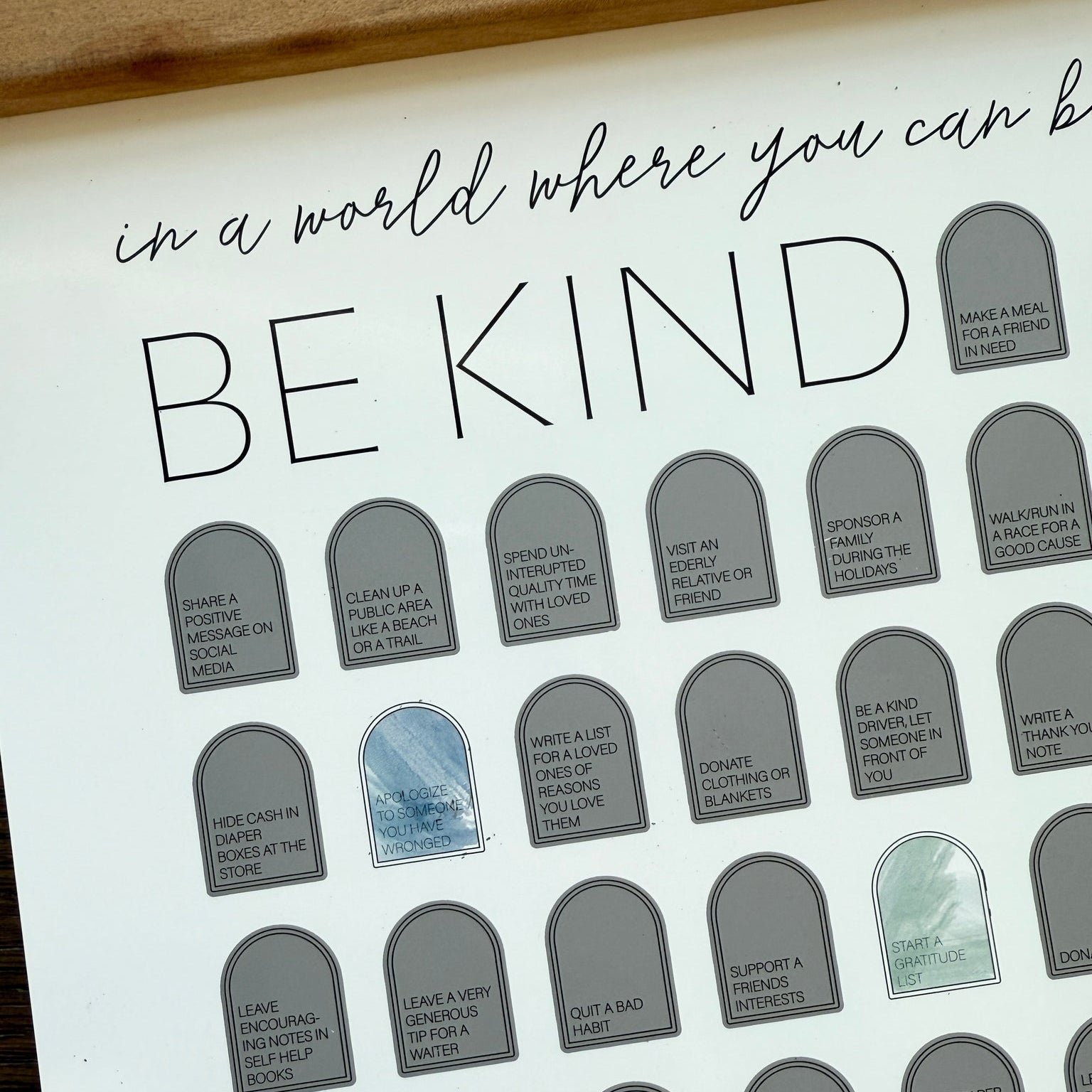 A scratch-off bucket list for acts of kindness. Some acts include, "Make a meal for a friend in need", "Sponsor a family during the holidays", and "Hide cash in diaper boxes at the store". "In a world where you can be anything, be kind" is written in script across the top. 