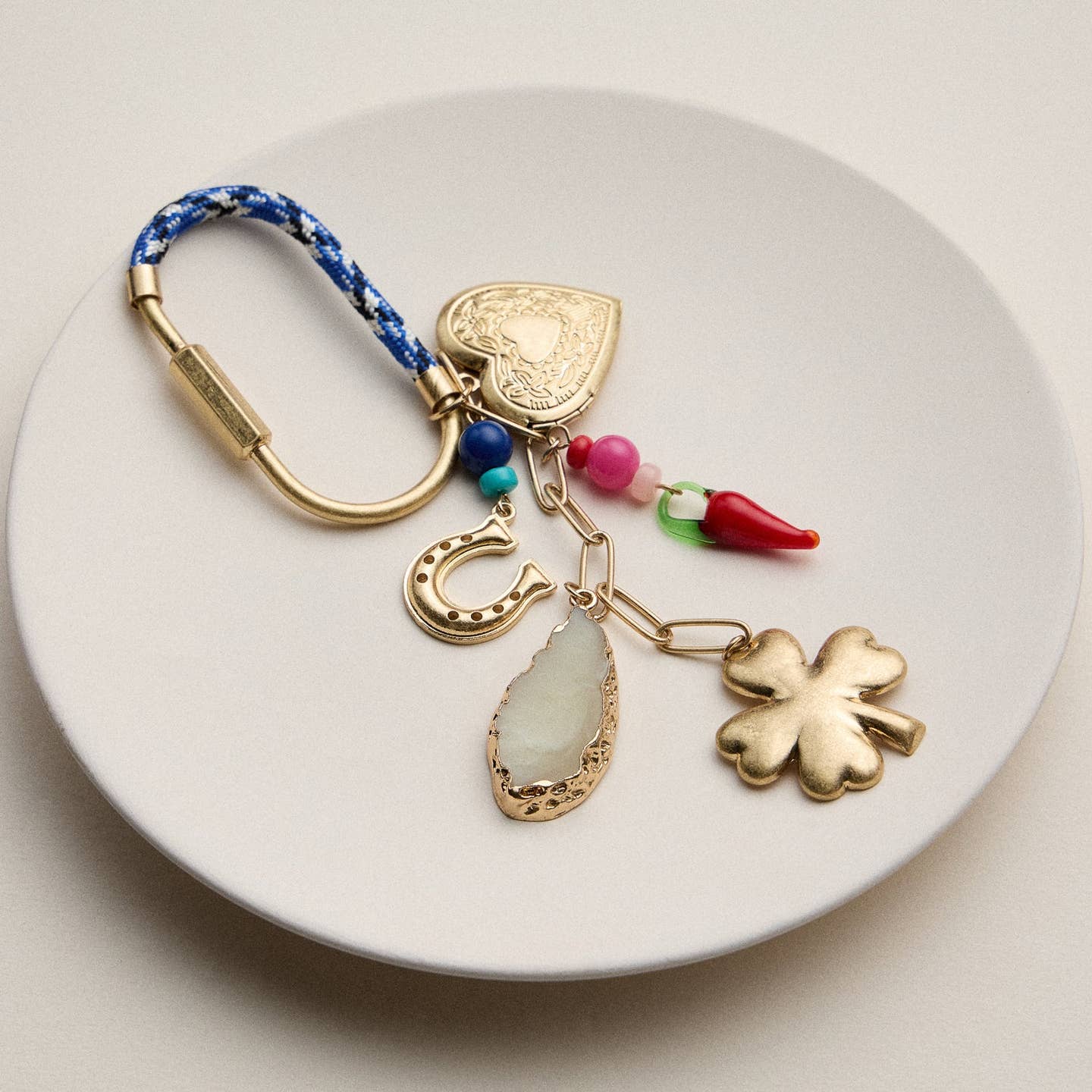 Carry a bit of good fortune wherever you go with this whimsical lucky charm keychain. Featuring a gold-tone heart locket, four-leaf clover, horseshoe, glass chili pepper, and a natural-style stone pendant, this eclectic accessory blends symbolism and style in one unique piece.

The beaded accents and blue woven loop give it a playful pop of color, while the gold metal details add a vintage-inspired touch. Use it as a keyring or clip it onto your tote, backpack, or purse for an eye-catching charm.