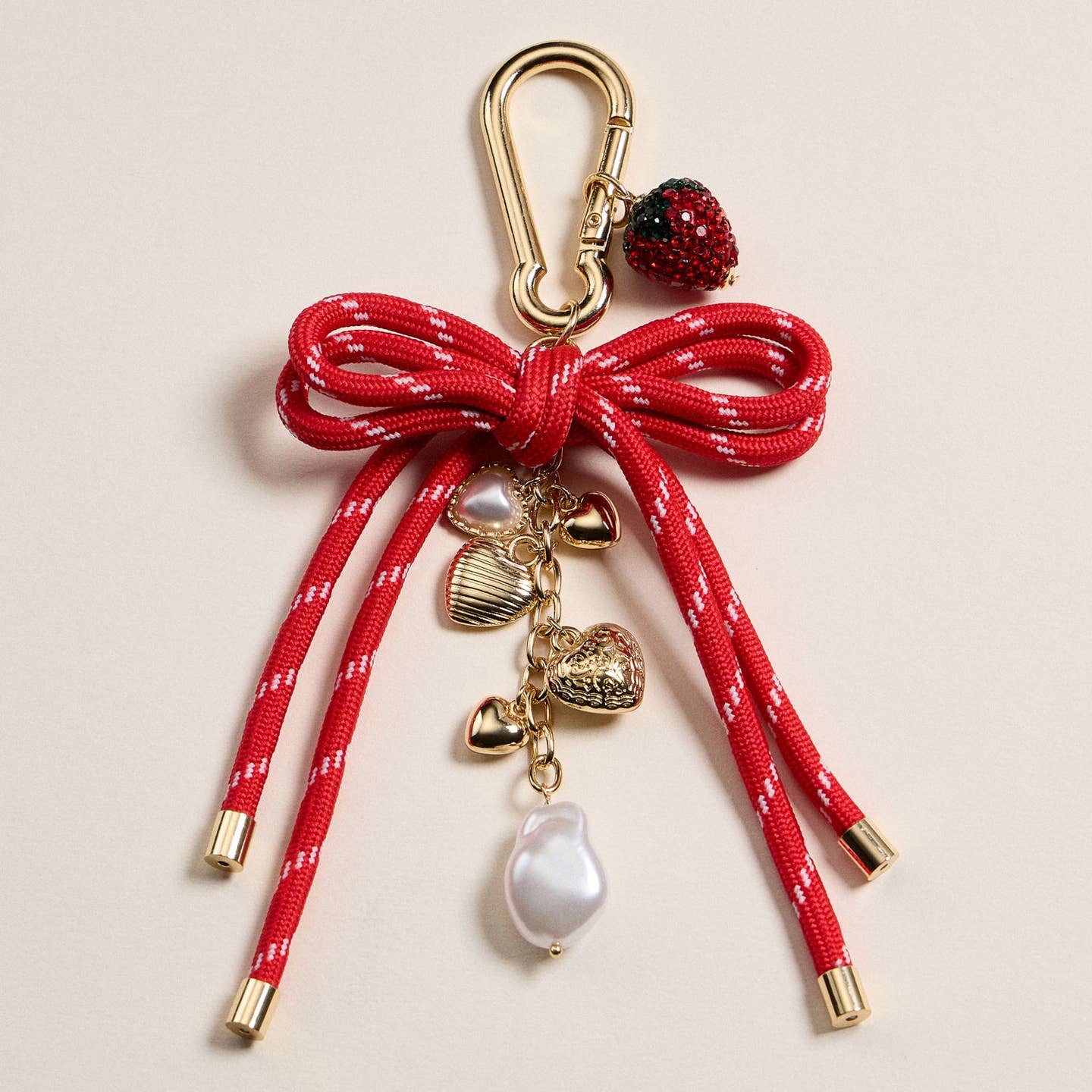 Add a sweet touch to your accessories with this enamel strawberry and heart charm keychain. Featuring a sparkling red strawberry, pearl accents, and multiple gold-tone hearts, all hanging from a vibrant red rope bow, this keychain brings charm and elegance to any bag or keyring. The sturdy gold-tone clasp ensures secure and easy attachment.

Product Features
Materials: Enamel charms, faux pearl, nylon rope, metal hardware
Charms: Strawberry, hearts, pearl accents
Length: Approx. 5.75 inches