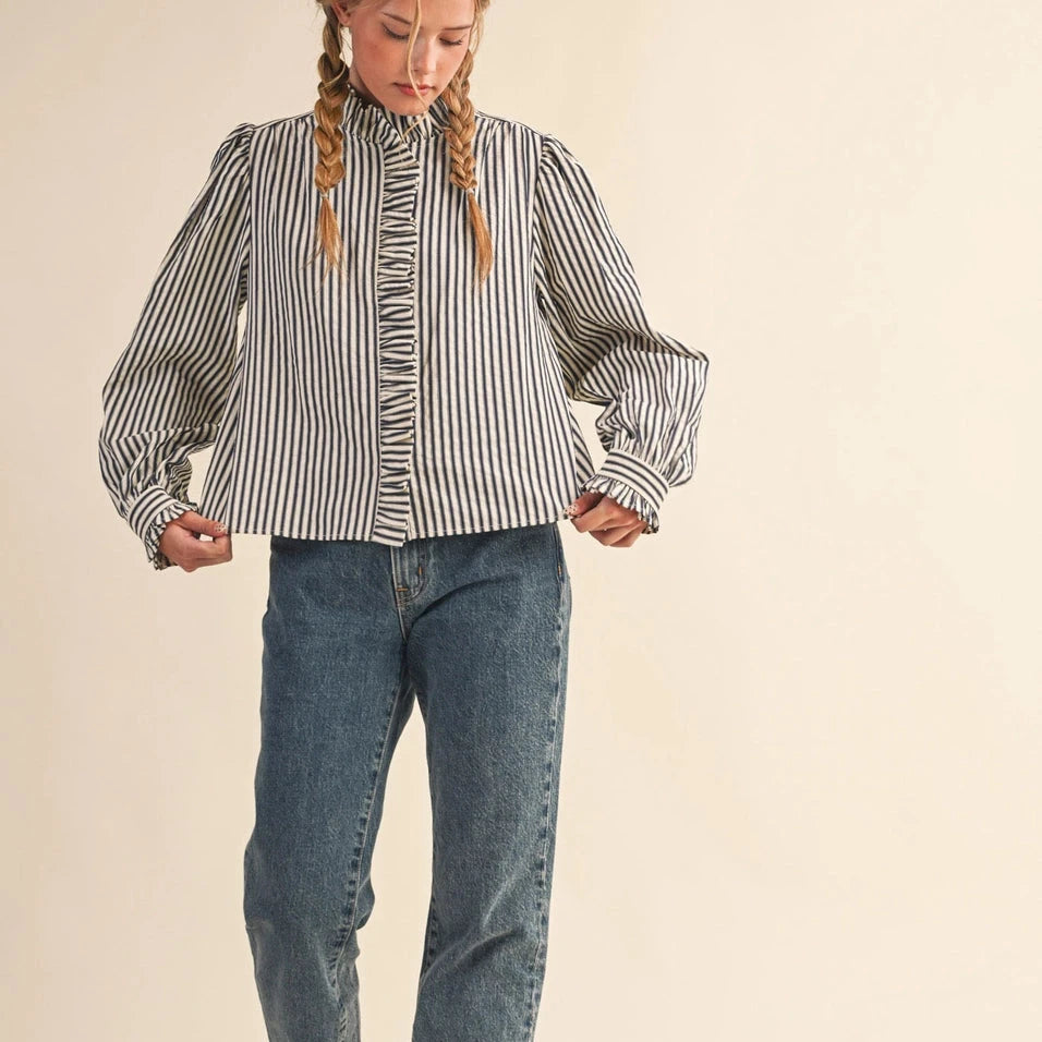 Embrace effortless elegance with this charming denim blue striped button down. Delicate ruffles frame the neckline and trim the placket, while puff sleeves with ruffled buttoned cuffs add a touch of vintage-inspired romance. The relaxed silhouette drapes beautifully, making it perfect for layering or wearing solo. Crafted from a comfortable polyester-cotton blend, this versatile top transitions seamlessly from casual weekends to polished occasions. A wardrobe essential.