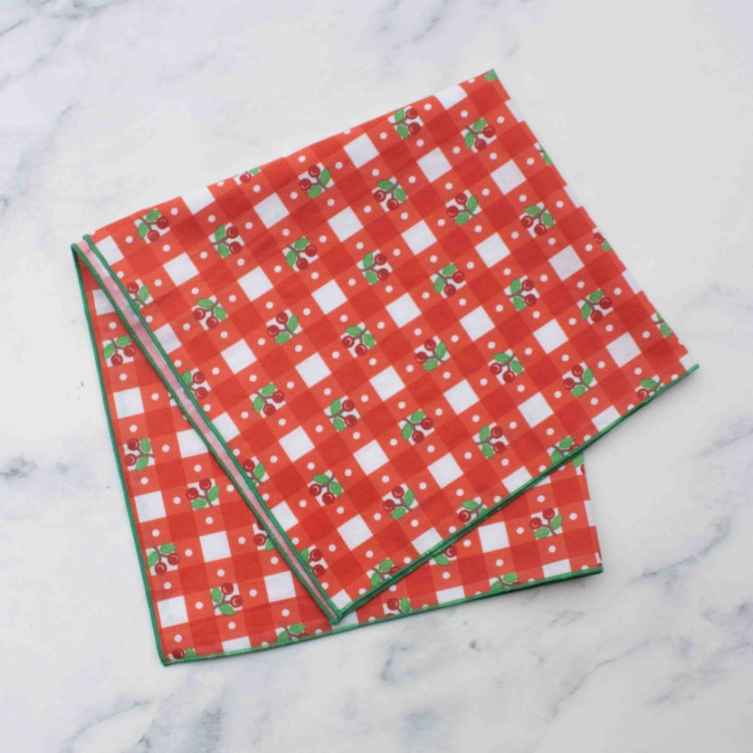Add a pop of playful charm to your look with our Cherry Gingham Square Scarf. Featuring a cheerful cherry motif over a classic gingham check, this lightweight scarf is made from 100% cotton for breathable comfort and everyday versatility. Its compact 17-inch (44cm) square size makes it perfect for tying around your neck, hair, or bag. Choose between vibrant red or refreshing blue – both finished with a colorful trim for a fun and polished touch.