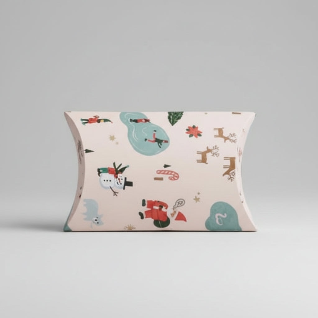 A Christmas-themed pillow box featuring a festive pattern with reindeer, Santa Claus, a snowman, and holiday motifs on a light cream background, set against a solid pale pink backdrop.