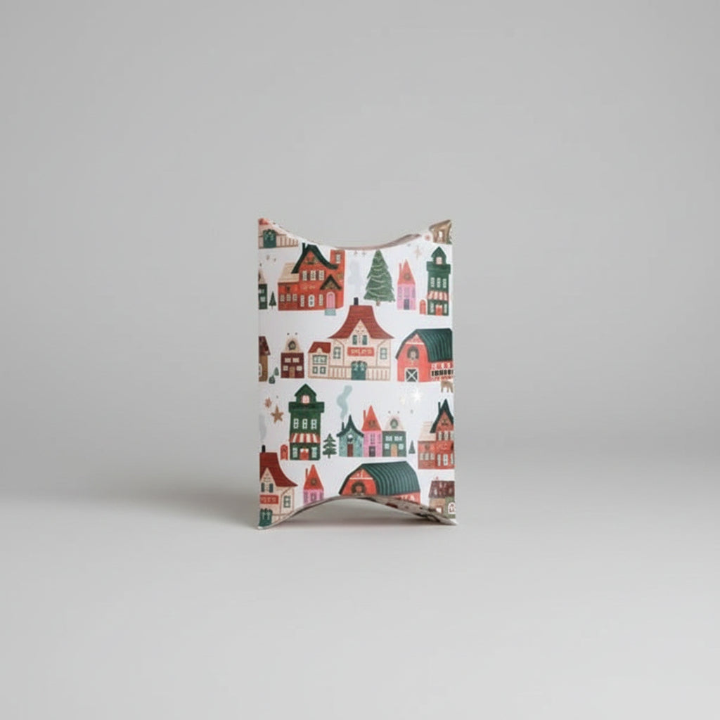 A square gift box with a festive winter village pattern featuring colorful houses, trees, and snow in red, green, blue, and yellow on a white background, set against a solid pale pink backdrop.