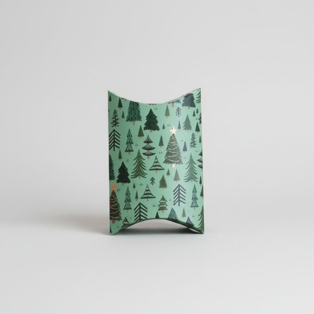 A gift box with a wavy top and bottom, featuring a repeating pattern of dark green pine trees on a mint green background, set against a solid pale pink backdrop.