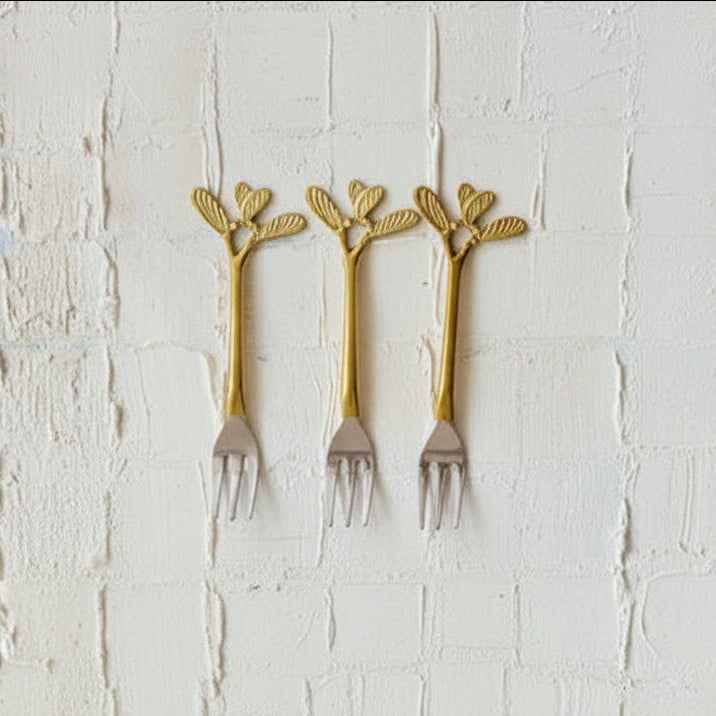 A flat-lay arrangement of a three-piece flatware set with decorative gold handles shaped like leaves or flowers, shown with matching fabric drawstring pouches labeled spoons, forks, and cutlery on a white brick background.
