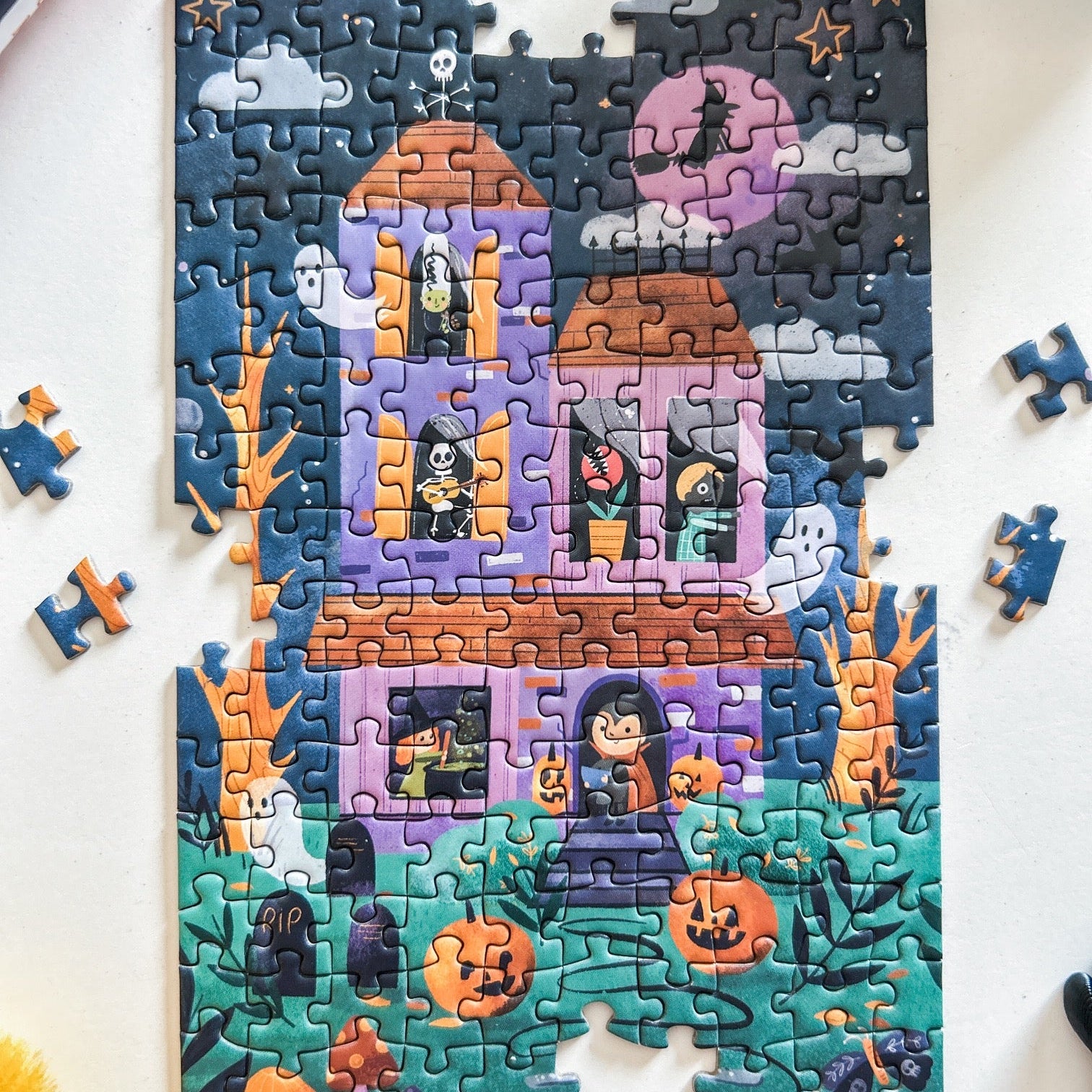 Mini Puzzle depicting a cute spooky mansion at night with jack-o-lanterns, ghosts, skeletons, zombies and witches. Pictured against a white back ground.